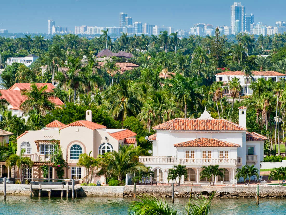 A picturesque waterfront view featuring luxurious homes with red tile roofs surrounded by lush palm trees, against a backdrop of a modern city skyline.